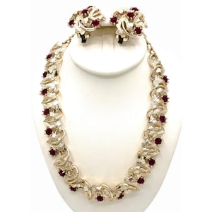 May include: A vintage jewelry set featuring a necklace and matching clip-on earrings. The set has a gold-tone metal finish with a leaf-like design. Each piece is adorned with small, round, red gemstones. The jewelry is displayed on a white bust.