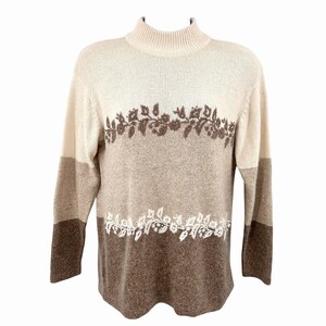 May include: A beige, brown, and tan sweater with a mock turtleneck. The sweater features a floral design in brown and white. The sleeves are long and the sweater is made of a soft material.