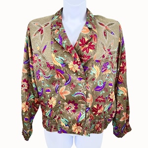 May include: A long-sleeve button-up blouse in a muted olive green base color featuring a vibrant floral print in shades of red, purple, yellow, and teal. The garment includes a notched collar, shoulder panels with a textured mesh overlay, and a gathered elastic waistband and cuffs.
