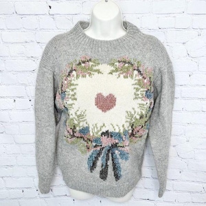 May include: A gray sweater with a floral heart design. The sweater features a crew neck and long sleeves. The heart design is in the center of the sweater and is surrounded by a wreath of flowers in shades of pink, green, and blue.