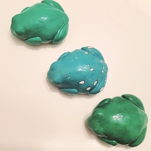 May include: Three ceramic frog figurines in varying shades of green and teal. One frog is teal with white spots, while the other two are solid green. The frogs are arranged in a diagonal line, showcasing their detailed, textured surfaces.