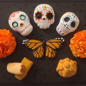 May include: A collection of Day of the Dead themed decorations. The arrangement includes three decorated sugar skulls, two orange paper flowers, a monarch butterfly, tamales, and a pan de muerto on a black background.