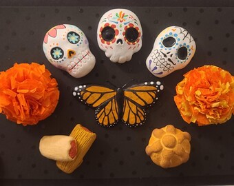 May include: A collection of Day of the Dead themed decorations. The arrangement includes three decorated sugar skulls, two orange paper flowers, a monarch butterfly, tamales, and a pan de muerto on a black background.