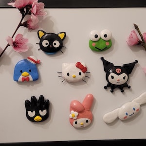 May include: A collection of colorful character magnets, including a black cat, a green frog, a blue penguin, Hello Kitty, a black bat-like character, a black bird, a pink bunny, and a white dog-like character. The magnets are arranged on a white surface, with pink flowers in the background.