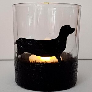 May include: A glass candle holder with a black silhouette of a dachshund dog. The candle holder has a black base and a clear glass top. A small candle is lit inside the holder.