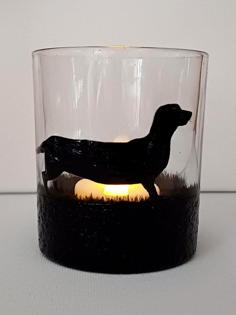May include: A glass candle holder with a black silhouette of a dachshund dog. The candle holder has a black base and a clear glass top. A small candle is lit inside the holder.