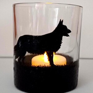May include: A clear glass candle holder with a black silhouette of a German Shepherd dog. The candle holder has a black base and a tea light candle is lit inside.