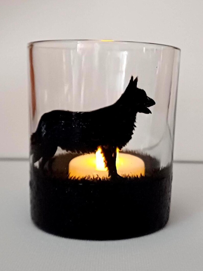 May include: A clear glass candle holder with a black silhouette of a German Shepherd dog. The candle holder has a black base and a tea light candle is lit inside.