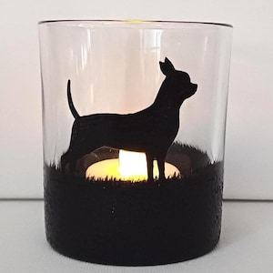 May include: A glass candle holder with a black silhouette of a Chihuahua dog standing in a field of grass. The candle holder has a black base and a clear glass top. A small tea light candle is lit inside the holder.