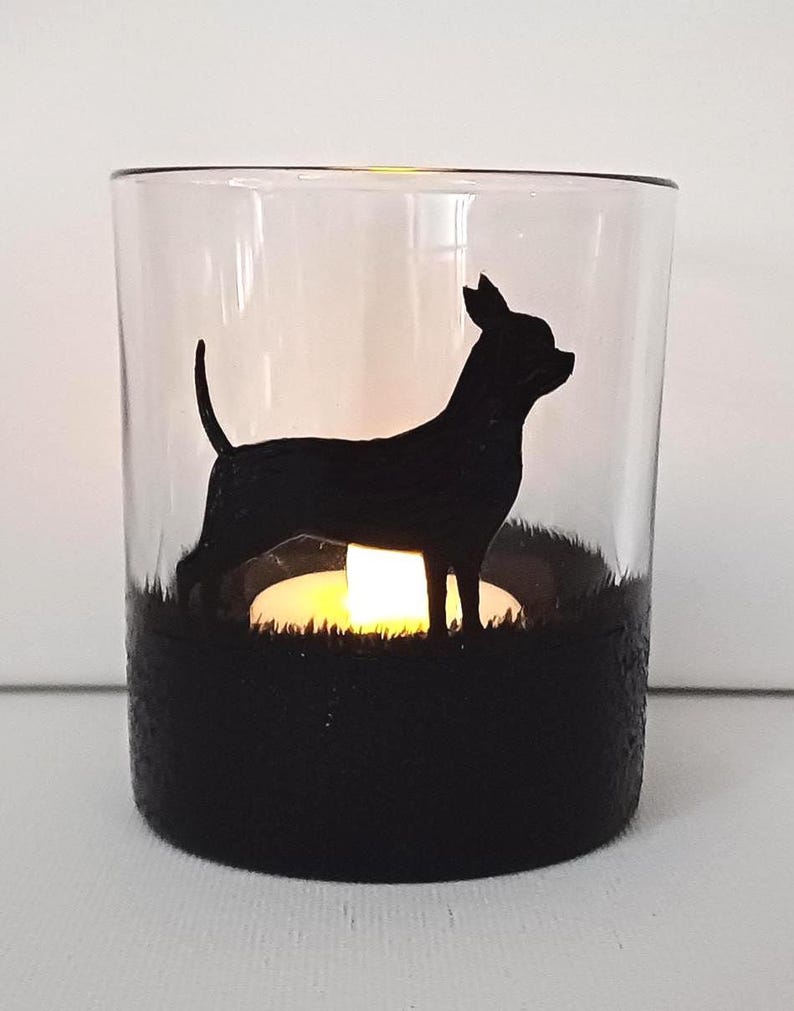 May include: A glass candle holder with a black silhouette of a Chihuahua dog standing in a field of grass. The candle holder has a black base and a clear glass top. A small tea light candle is lit inside the holder.