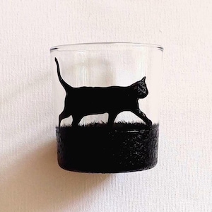 May include: A clear glass tumbler featuring a black cat silhouette walking on a grassy base. The bottom section of the glass is painted black, providing a textured appearance. The cat's form is clearly defined, capturing its movement.