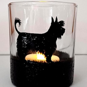 May include: A glass candle holder with a black silhouette of a dog. The candle holder has a black base and a clear glass top. A small white candle is lit inside the holder.
