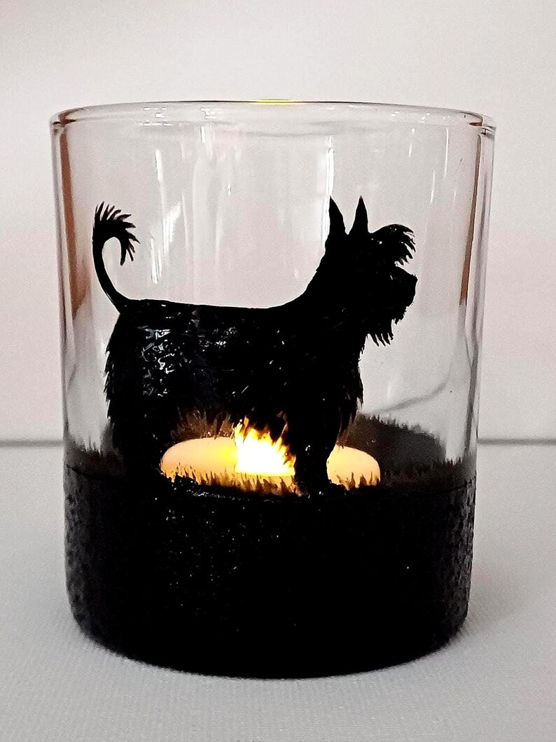 May include: A glass candle holder with a black silhouette of a dog. The candle holder has a black base and a clear glass top. A small white candle is lit inside the holder.