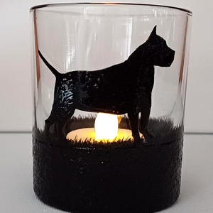 May include: A glass candle holder with a black silhouette of a dog standing in a field of grass. The candle holder has a black base and a clear glass top. A small white tea light is lit inside the holder.
