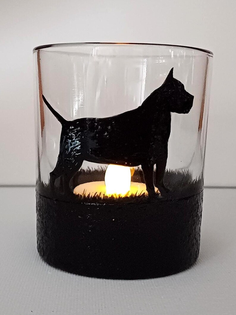 May include: A glass candle holder with a black silhouette of a dog standing in a field of grass. The candle holder has a black base and a clear glass top. A small white tea light is lit inside the holder.