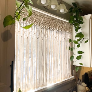 May include: A white macrame curtain hangs in a window with a white trim. The curtain is made of knotted cords and has a fringe at the bottom. There are two houseplants on either side of the window.