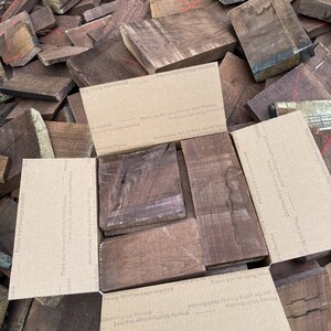 May include: A pile of dark brown wood planks, some with a reddish hue, are stacked on top of each other. The wood is arranged in a random pattern. The wood is likely for woodworking projects. The wood is surrounded by brown cardboard boxes with text that says "Priority Mail postage required" and "Thank you for using Priority Mail Service".