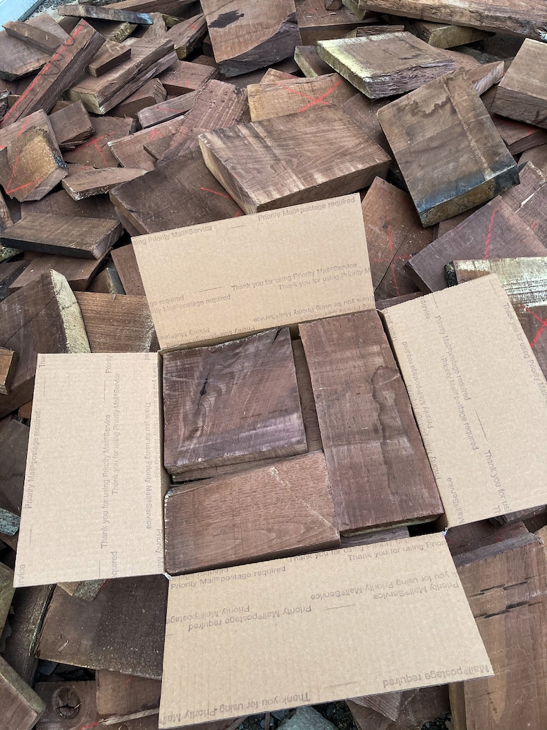 May include: A pile of dark brown wood planks, some with a reddish hue, are stacked on top of each other. The wood is arranged in a random pattern. The wood is likely for woodworking projects. The wood is surrounded by brown cardboard boxes with text that says "Priority Mail postage required" and "Thank you for using Priority Mail Service".