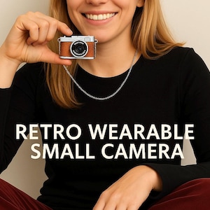Puede incluir: Un pequeño colgante con forma de cámara retro, con un acabado marrón efecto cuero y detalles plateados. La cámara tiene una lente negra y cuelga de una cadena plateada. El texto "RETRO WEARABLE SMALL CAMERA" se muestra debajo.