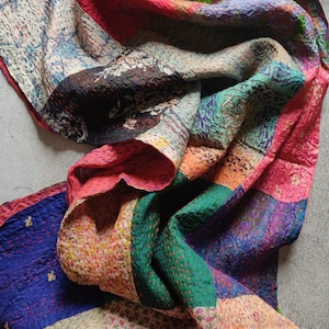 May include: A vibrant patchwork textile in shades of red, green, blue, and pink. The fabric is composed of various patterned pieces stitched together, with visible seams. The design is visually striking, featuring a mix of floral and abstract patterns.
