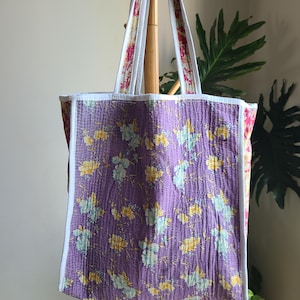 May include: A quilted tote bag with a floral pattern in shades of purple, yellow, and blue. The bag has white trim and handles with a floral design. The tote is hanging on a wooden rack, showcasing its design and construction.