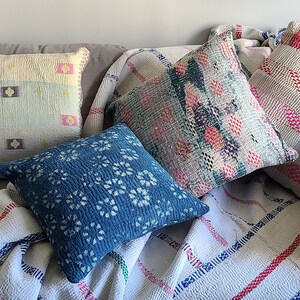 Peut inclure: Cinq coussins de décoration à motifs, dans des couleurs et des designs variés, dont le rose, le bleu et le blanc, disposés sur un canapé gris avec une couverture en laine rayée blanche et rouge.