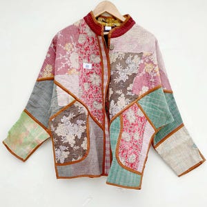 May include: A long-sleeve quilted jacket featuring a patchwork design of various floral and textured fabrics in shades of pink, green, brown, and cream. The jacket has a mandarin collar, a front button closure, and brown trim along the seams.