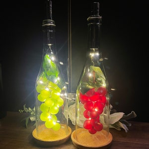 May include: Two glass bottles with cork tops, filled with grapes and fairy lights. The bottle on the left has green grapes and the bottle on the right has red grapes.