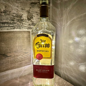 Puede incluir: Una botella de vidrio transparente con una etiqueta dorada que dice "Jose Cuervo Especial" y "Made with Blue Agave Gold Tequila". La botella está decorada con luces blancas.