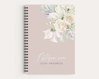 May include: A spiral bound notebook with a light pink cover featuring a watercolor illustration of white flowers and greenery. The text "Notizen von LUISA FRIEDRICH" is printed in white on the cover.