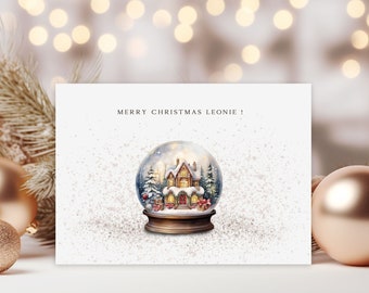 May include: A Christmas card with the text "MERRY CHRISTMAS LEONIE!" features a snow globe illustration. The snow globe showcases a cozy house scene with trees and presents. The card is set against a blurred background with gold ornaments.