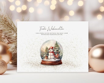 May include: A Christmas card with a snow globe illustration featuring a snowman, trees, and gifts. The card has the text "Frohe Weihnachten" and a holiday greeting. Gold ornaments and a sprig of greenery are in the background.