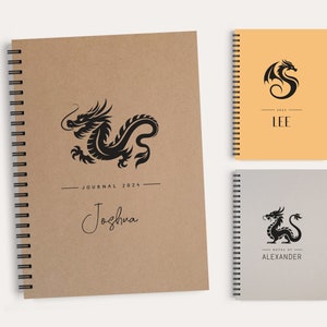 Könnte beinhalten: Spiralgebundene Notizbücher mit Drachendesigns. Das größte Notizbuch ist braun mit einer schwarzen Drachenillustration, dem Text "JOURNAL 2024" und dem Namen "Joshua". Zwei kleinere Notizbücher sind ebenfalls abgebildet, eines orangefarbenes und eines graues, jeweils mit einem Drachenlogo.