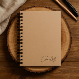 Könnte beinhalten: Ein spiralgebundenes Notizbuch mit einem schlichten hellbraunen Einband, auf dem der Name Charlotte in schwarzer Schreibschrift in der unteren rechten Ecke aufgedruckt ist. Das Notizbuch hat eine schwarze Metallspirale an der linken Seite.