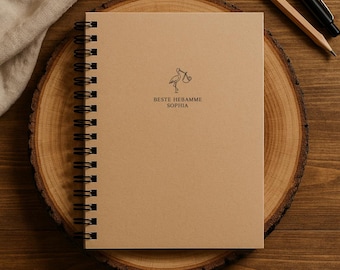 May include: A spiral-bound notebook with a kraft paper cover. The cover features a stork illustration carrying a baby and the text "BESTE HEBAMME SOPHIA" in black. The notebook rests on a wooden surface with a pencil and pen.