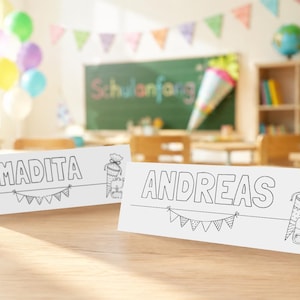 Puede incluir: Dos tarjetas de nombre de papel blanco plegado con diseños de arte lineal en negro. Una tarjeta presenta el nombre MADITA y la otra el nombre ANDREAS. Ambas tarjetas muestran una guirnalda de banderines triangulares y un cono escolar.
