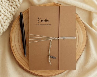 May include: A brown journal with the name "Emelies" and the text "GEDANKENSAMMLER" printed on the cover. The journal is tied with white string and a small feather charm. A black pen rests beside it on a wooden slice.