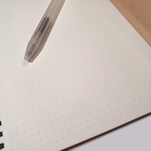 May include: A spiral-bound notebook with off-white dot grid paper and a brown cover, featuring a clear plastic ballpoint pen resting on the open page. The notebook has a black metal wire binding along the left side, providing a clean surface.