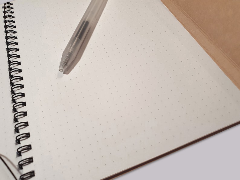 May include: A spiral-bound notebook with off-white dot grid paper and a brown cover, featuring a clear plastic ballpoint pen resting on the open page. The notebook has a black metal wire binding along the left side, providing a clean surface.