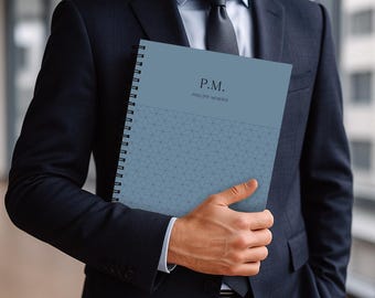 May include: A blue spiral-bound notebook with the initials "P.M." and the name "PHILIP MORRIS" printed on the cover. The notebook has a geometric pattern on the lower half of the cover and is held by a person wearing a dark suit.