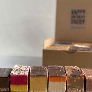 Op de afbeelding: Een doos met verschillende fudge-vierkanten in smaken zoals chocolade, vanille en karamel. De fudge is in een rij gerangschikt, waarbij elk vierkant een andere smaak en kleur heeft.