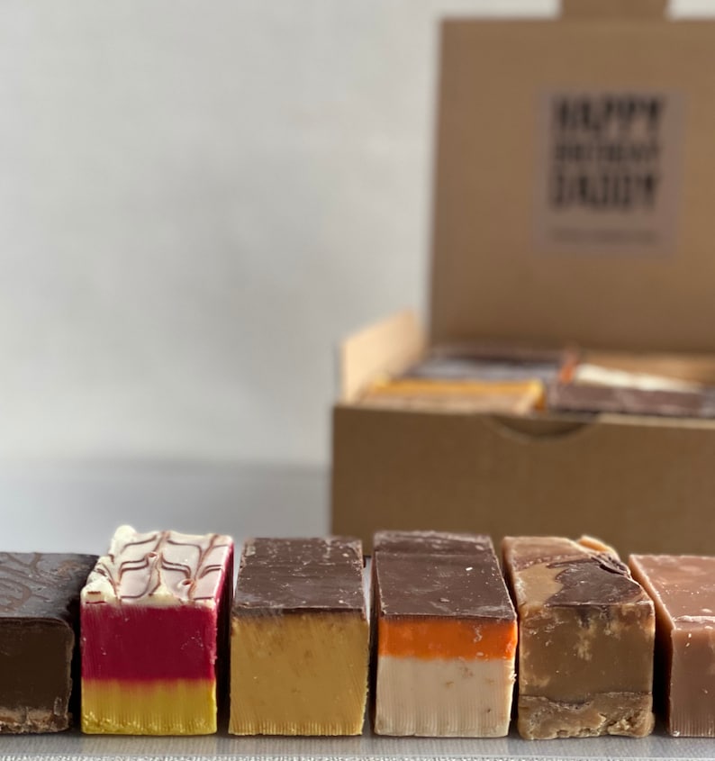 Op de afbeelding: Een doos met verschillende fudge-vierkanten in smaken zoals chocolade, vanille en karamel. De fudge is in een rij gerangschikt, waarbij elk vierkant een andere smaak en kleur heeft.