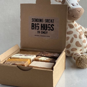 Op de afbeelding: Een bruine kartonnen doos met een open deksel, waarin verschillende fudge-vierkanten te zien zijn. De doos heeft een sticker met de tekst "Sending Great Big Hugs To: Emily Original Handmade Fudge". Een knuffelgiraf zit naast de doos.