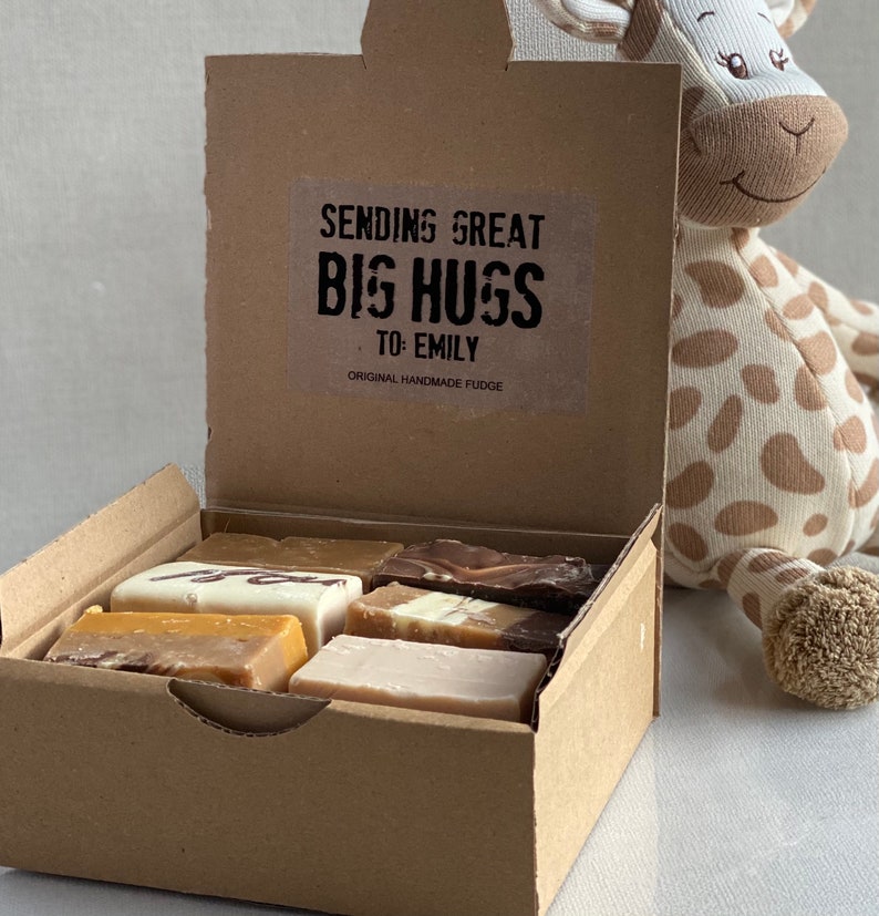 Op de afbeelding: Een bruine kartonnen doos met een open deksel, waarin verschillende fudge-vierkanten te zien zijn. De doos heeft een sticker met de tekst "Sending Great Big Hugs To: Emily Original Handmade Fudge". Een knuffelgiraf zit naast de doos.