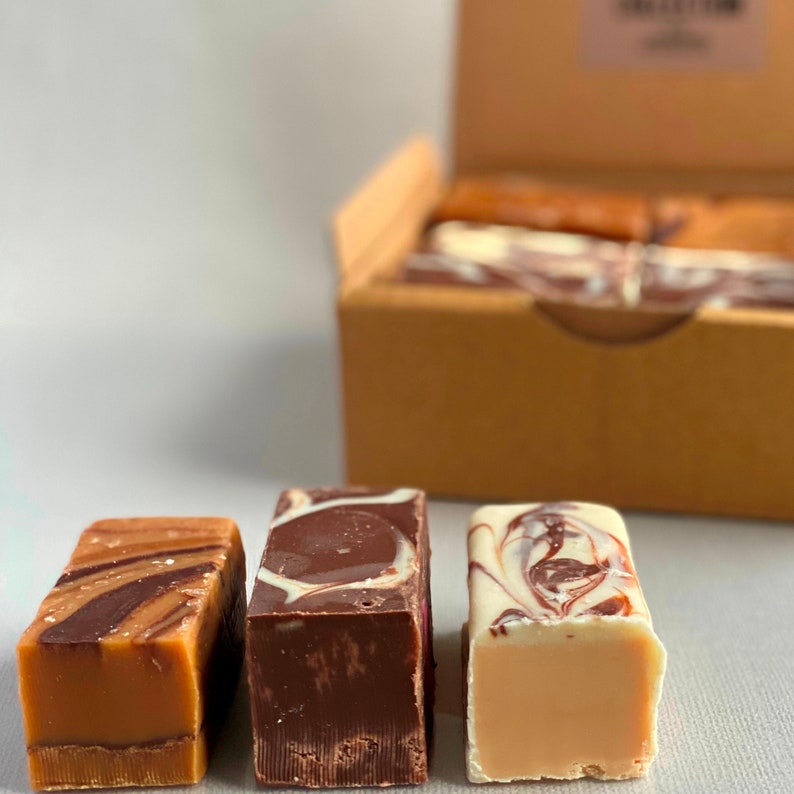 Op de afbeelding: Drie verschillende smaken fudge, elk met een swirl-patroon. De fudge is in een rij op een wit oppervlak gerangschikt. De fudge-smaken zijn karamel swirl, chocolade swirl en vanille swirl.