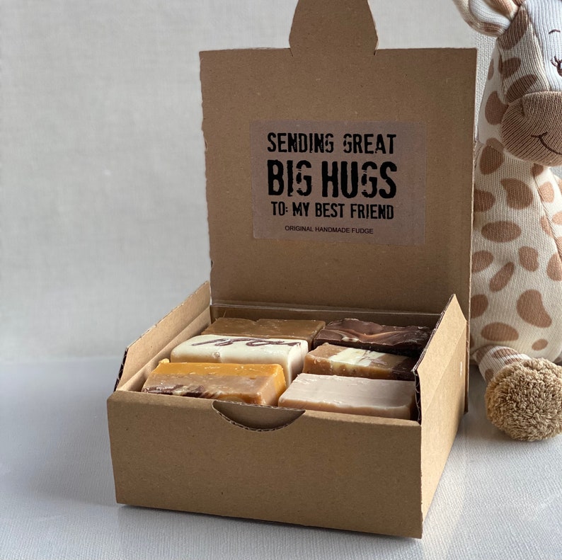 Op de afbeelding: Een bruine kartonnen doos met een open deksel, waarin verschillende fudge-vierkanten te zien zijn. De doos heeft een sticker op het deksel met de tekst "Sending Great Big Hugs To My Best Friend Original Handmade Fudge". Rechts in de afbeelding staat een knuffeldier in de vorm van een giraffe.