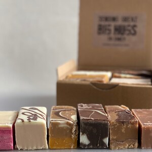 Op de afbeelding: Een doos met verschillende fudge-soorten met smaken zoals chocolade, vanille en karamel. De doos is gelabeld met "Scrumptious Scrummy Big Mess".