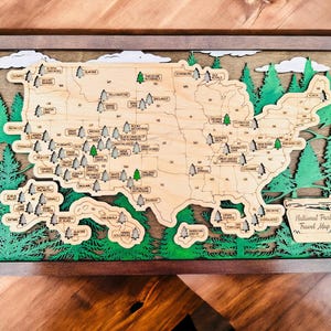 May include: Wooden National Park Travel Map of the United States, framed in gray. The map is layered with a light wood base, green trees, and white clouds. Each park is labeled with a small tree icon.