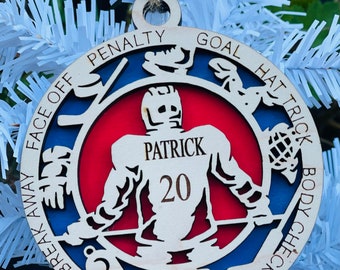 Può includere: Un ornamento di hockey in legno con un design rosso, bianco e blu. L'ornamento presenta un giocatore di hockey al centro con il nome "Patrick" e il numero "20" sulla maglia. L'ornamento presenta anche vari termini di hockey attorno al bordo, tra cui "penalità", "gol", "hat trick", "corpo a corpo", "fuga", "faccia a faccia", "tiro di barra" e "finta".