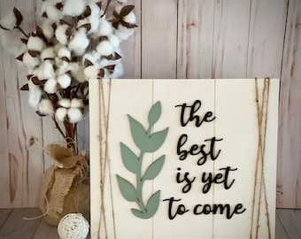 May include: A wooden sign with the phrase "The best is yet to come" in black script, accompanied by a light green leaf design. The sign is decorated with twine and set against a rustic wooden background. A cotton plant in a burlap bag is to the left.